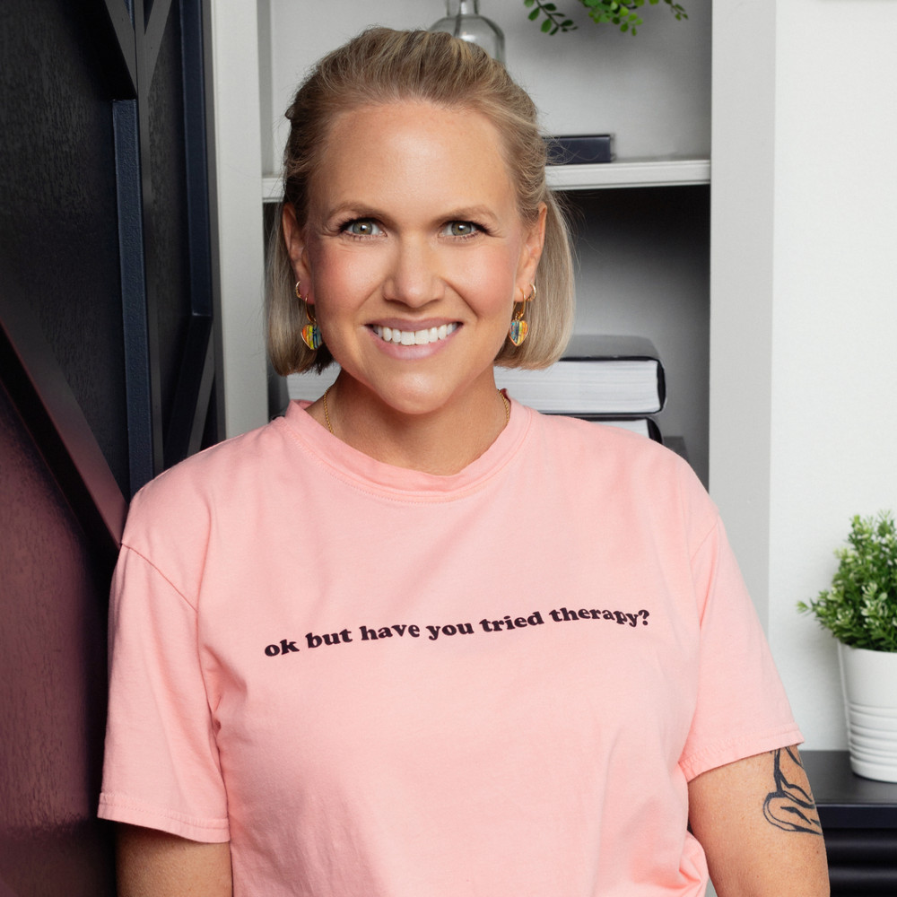 SarahBeth Bolin, LCSW, smiling at the camera in a pink t-shirt printed with the words “ok but have you tried therapy?”, wearing rainbow heart earrings.